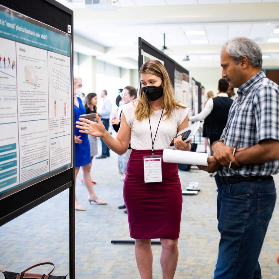 A female trainee explains her research poster to male attendee at research day.