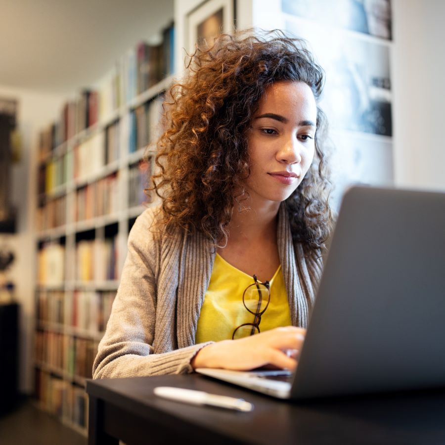 A woman with dark curly hair sits in front of a laptap doing work.