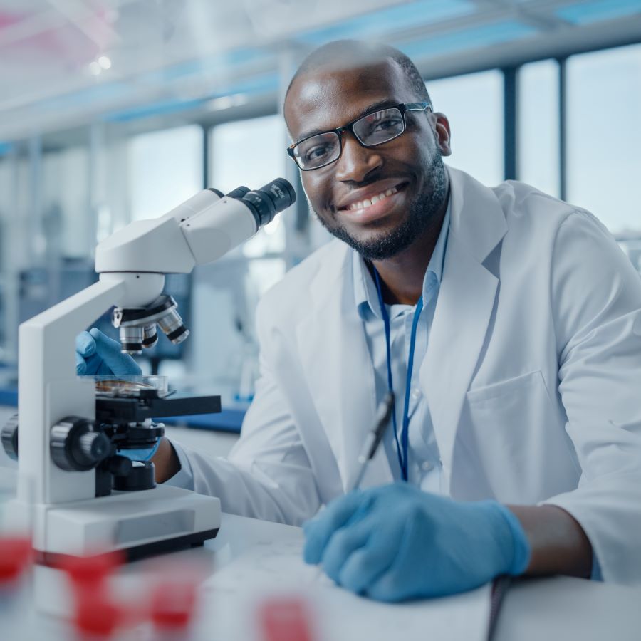 A Black male researcher wearing glasses and a lab coat takes notes while looking at a microscope.