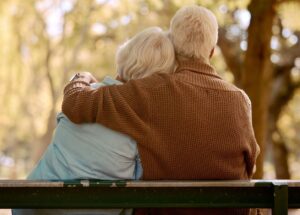 An older man and woman sit on a bench facing away from the camera. The man has his arm around the woman's shoulders.